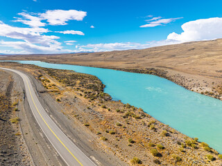 Aerial view of a bright turquoise glacial river flowing alongside a paved highway through a scenic arid landscape under a blue sky