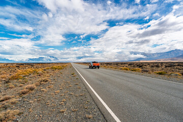 Orange pickup truck driving on a straight asphalt road through the vast wilderness of Patagonia, Argentina, with mountains and clouds in the background