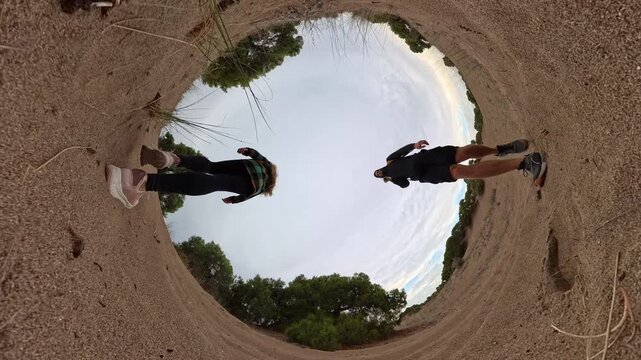 A "tiny planet" perspective captures a man and a woman walking through a sandy landscape surrounded by sparse trees. The spherical view emphasizes their steps and the expansive environment