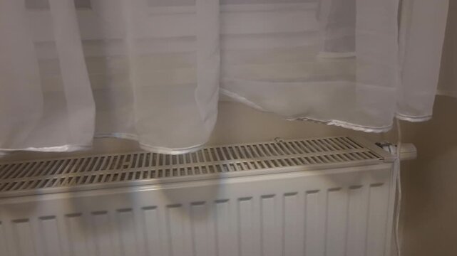 Close-up of light curtains hanging above radiator with camera slowly rotating to the right. Soft daylight highlights fabric texture and creates peaceful indoor home environment.