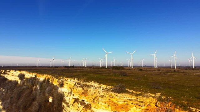 Wide aerial view of multiple wind turbines scattered across a green and brown field adjacent to a rocky sea cliff. Wind farm on field near coastal line.