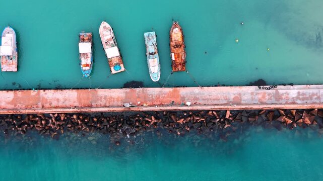 Aerial view of fishing boats docked at a concrete pier with tetrapods in turquoise water. Top down view of boats moored at pier.