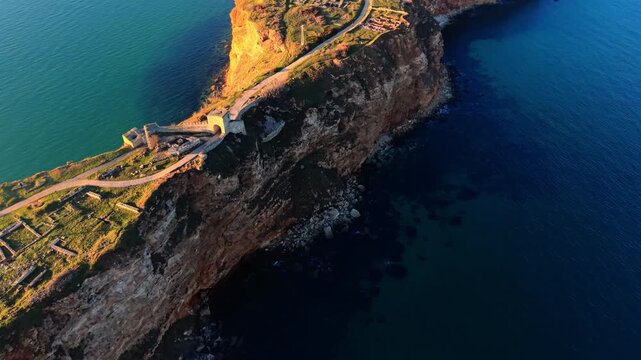 Aerial sunset view of Cape Kaliakra with medieval walls and steep rocky cliffs. Ancient fortress ruins on a narrow cliff peninsula.