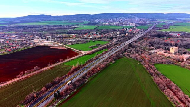 An aerial perspective of a multilane motorway stretching through green and brown fields near a small town and hills. Long straight highway passing through agricultural landscape.