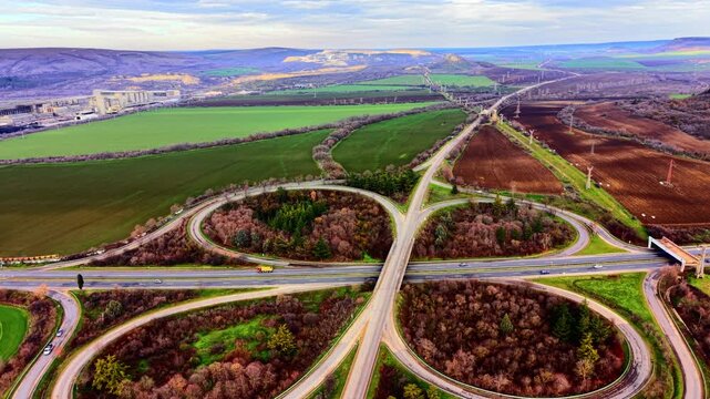 A complex road junction with multiple ramps surrounded by agricultural fields and industrial buildings in the distance. Aerial view of a highway cloverleaf interchange in countryside.