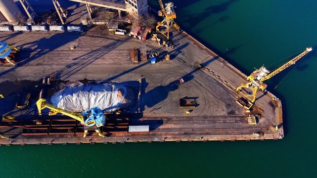 Industrial cranes, shadows on the ground, and a bulk cargo pile covered with a grey tarp. Top down aerial view of yellow port cranes on concrete pier.