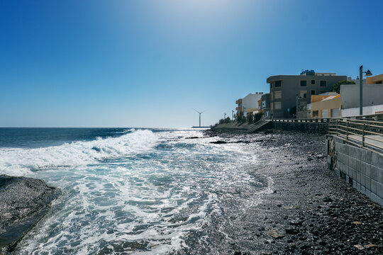 A coastal view of Arinaga on Gran Canaria, showing waves breaking against the rocky shoreline beside a modern seaside promenade. Residential buildings