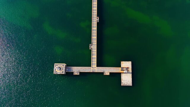 Modern pier structure with a lighthouse at the end, surrounded by deep green sea water. T shaped concrete pier. Aerial view.