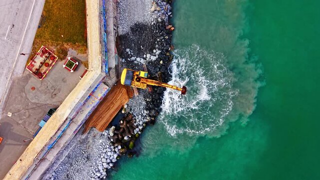Yellow excavator placing stones on a sea wall, creating sediment plumes in the clear green water. Aerial top down shot of excavator moving rocks on coast.