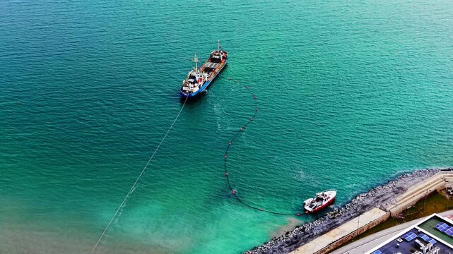 Larger red ship and a smaller boat managing a long floating pipe connected to the shore. Aerial view of dredging ships and pipeline near coastline.