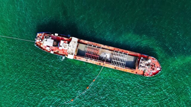 Red ship deck and open hold with a floating discharge pipe in rippling green sea. Close up top view of industrial dredging vessel in water.