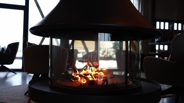 Suspended cone hood glass fireplace with flickering logs anchors a modern lounge in an Alps chalet, with snowy peaks through floor to ceiling windows at daytime.