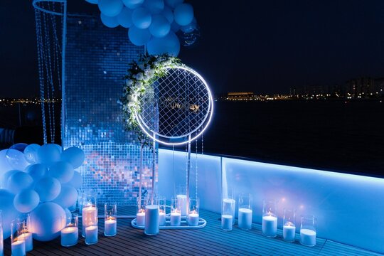 Nighttime outdoor wedding deck decoration illuminated by blue neon lights, featuring a glowing floral arch, blue balloons, and lit candles in glass vases.