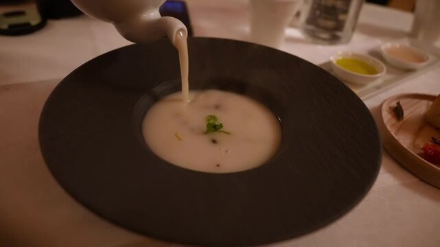 Close up shows a slow pour of steaming creamy soup into a textured black bowl, garnished with microgreens and capers, with olive oil, pink salt, and bread board nearby.