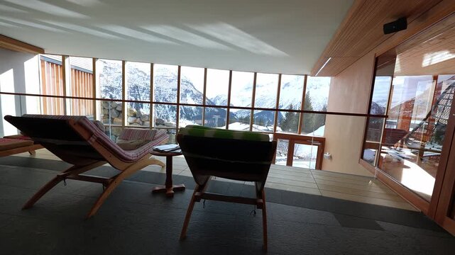 Interior spa mezzanine shows two wooden recliners and side table by mullioned windows, glass balustrade, and snow covered Alps with nearby chalet rooftops.