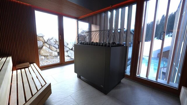 Camera pans through wood paneled sauna with slatted benches, matte black stove, arched guards, and windows to Alpine snow, rock wall, conifers, and heated pool.