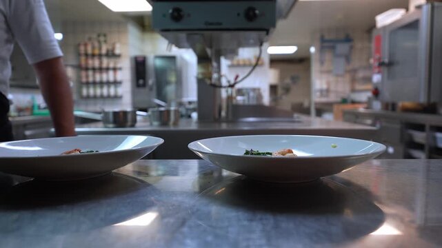 Two white plates sit on the pass as a chef in black apron reaches to garnish. Soft overhead light, shallow depth of field, bokeh, ovens and spice rack suggest Alps hotel kitchen.