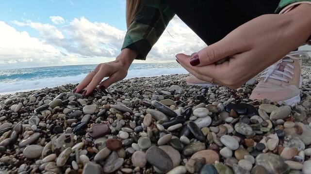 Close-up view of a woman's hand collecting colorful pebbles on a beach, with waves and a dramatic cloudy sky in the background