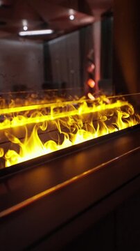Vertical shot shows a linear gas fireplace with glass, bright flames, and mirrored reflections, with lounge seating and tall curtains in soft focus at night.