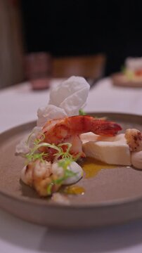 Tight vertical view shows char grilled prawn on white mousse, seared bites, puffed crisps, microgreens, and golden sauce on stoneware, slow tilt in warm light.
