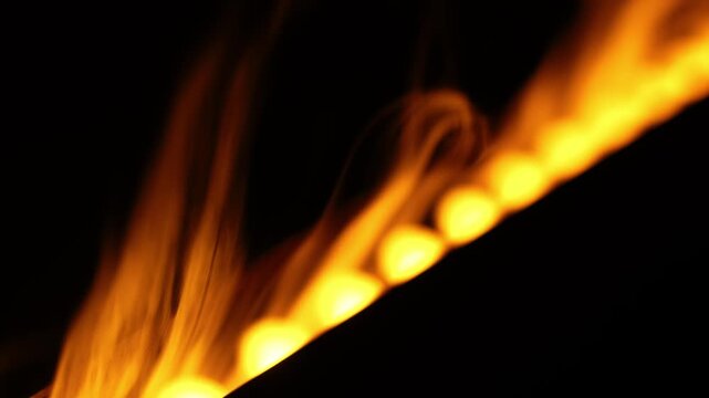 Evenly spaced golden flames flicker and stretch upward on a dark indoor set, forming a linear burner effect with shallow depth, rich amber tones, and high contrast.