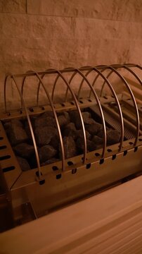 Close view of a sauna heater with black stones and a curved steel safety cage. The camera slowly pans under warm amber light, vertical frame, indoor spa mood.