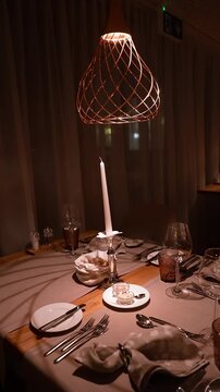 An intimate dining table pans and tilts under a copper wire pendant as a candle and two ramekins flicker. Wine glasses, linens, and an exit sign set an alpine hotel mood.