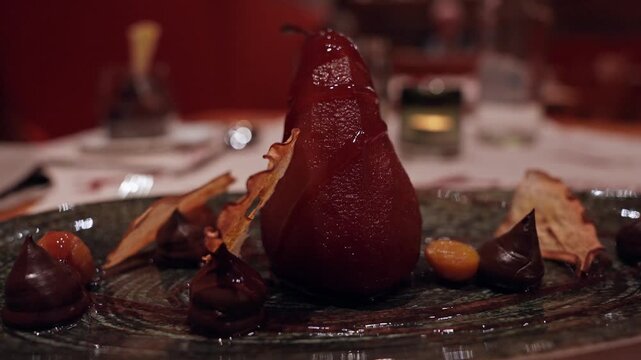 Glossy poached pear stands on a textured dark plate with ganache, candied apricot, and fruit chips, white tablecloth and candle blur, slow lateral camera move