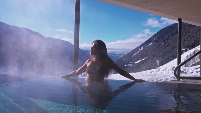 A woman in a bikini leans on a steaming outdoor infinity pool at a modern chalet, framed by concrete and metal, with Alpine slopes and a deep valley in winter daylight.
