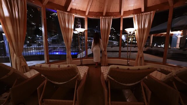 A white robed person pulls curtains in an octagonal wooden spa rotunda, wicker loungers and brass lamps glow warm, cool purple pool lights and snowy terrace beyond.