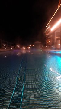 Low angle night view of a heated infinity pool with mosaic floor, blue, green and purple LED light, steam, lounge chairs, and a lit hotel terrace in Ischgl, Austria.