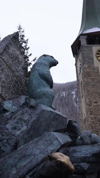 Low angle outdoor view shows bronze marmot sculptures on carved rock before a stone church tower with pointed green spire and clock faces in Zermatt, winter pans