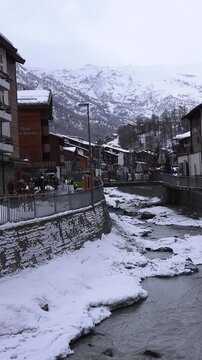 Vertical winter scene in Zermatt, Switzerland, with a narrow river, Haus zur Bruecke sign, wooden chalets, pedestrians, shuttle buses, and overcast cool light.