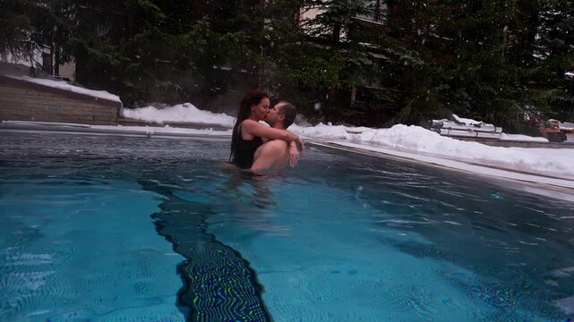 A couple in swimwear embrace and move in a heated infinity pool in Zermatt, Switzerland, low angle, snow falling, steam rising, ripples spread, overcast winter light.