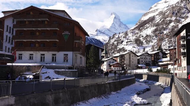 Sunlit winter street in Zermatt shows Hotel Perren, chalets, church steeples, and a snow lined river as people stroll, with the Matterhorn rising in the background.