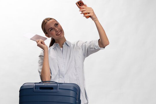 Smiling woman with long hair holding passport and smartphone takes selfie near blue suitcase. Concept of travel, vacation, tourism, and modern lifestyle with happy mood and casual outfit.