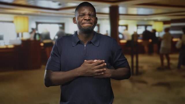 Man clutching hands over chest, grimacing at front desk in building reception with blurred people, counter and lamp visible; pain emergency.