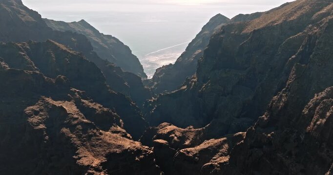 Masca Valley with mountain canyon leading to the glistening Atlantic Ocean, volcanic cliffs framing a boat wake amid dramatic coastal scenery on Tenerife, Canary Islands. Aerial view drone flight