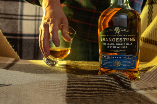 Close-up of a hand holding a filled glencairn glass next to a Grangestone Whisky bottle on a checkered plaid. Cozy lifestyle with soft natural lighting. Ukraine, Zhytomyr, March 29, 2026