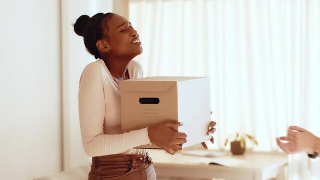 A woman with dark skin stands in a well-lit room, smiling as she receives a box from another person. The atmosphere is friendly and welcoming, suggesting a moment of connection.