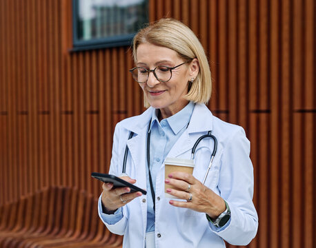 Portrait of a mature older female woman doctor or nurse and  health care worker using a smartphone mobile phone  in front of hospital or clinic outside
