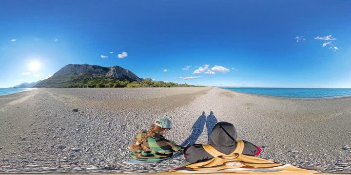 360-degree panorama of a couple walking along a pebbled beach, casting long shadows under the bright sun, with turquoise water and green cliffs in the background