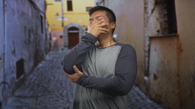 Man covers mouth with hand on cobblestone street by a yellow building and arched doorway; shy amusement.