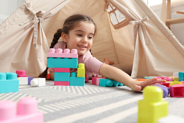 Little girl playing with building blocks near toy wigwam in playroom, low angle view © New Africa