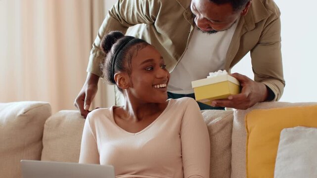 A Black woman sits on a couch using a laptop while a man with dark skin playfully covers her eyes. He holds a small gift box, ready to surprise her during a special occasion.