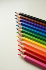 Fototapeta premium Rainbow colored pencils arranged diagonally displaying full spectrum from black through blue, green, yellow, orange, red to pink on neutral background.