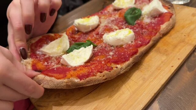 rectangular Italian pizza with tomato, cheese, and basil. Woman with brown manicure pulls apart a sliced piece. Rustic street food scene in Italy, fresh and appetizing.