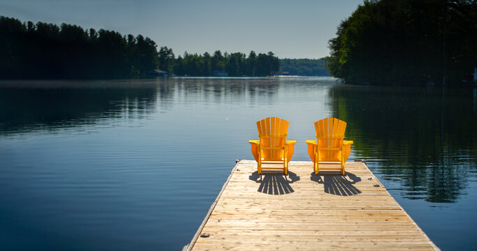 Rear view of two yellow Adirondack chairs on a dock at sunrise in Muskoka Ontario Canada, long shadows on calm glassy lake, cottage country, no people.
