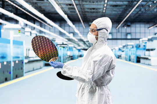 Man in bunny suit evaluating silicon wafer in brightly lit manufacturing plant cleanroom. Engineer checking semiconductor substrate nanometer circuit in modern foundry facility