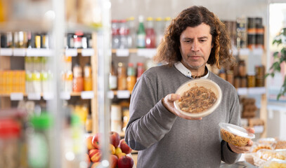 Fototapeta premium Adult man holds a package of ready-made yakisoba noodles, reading the ingredients and looking at the expiration date. European man buys cooked food in a supermarket